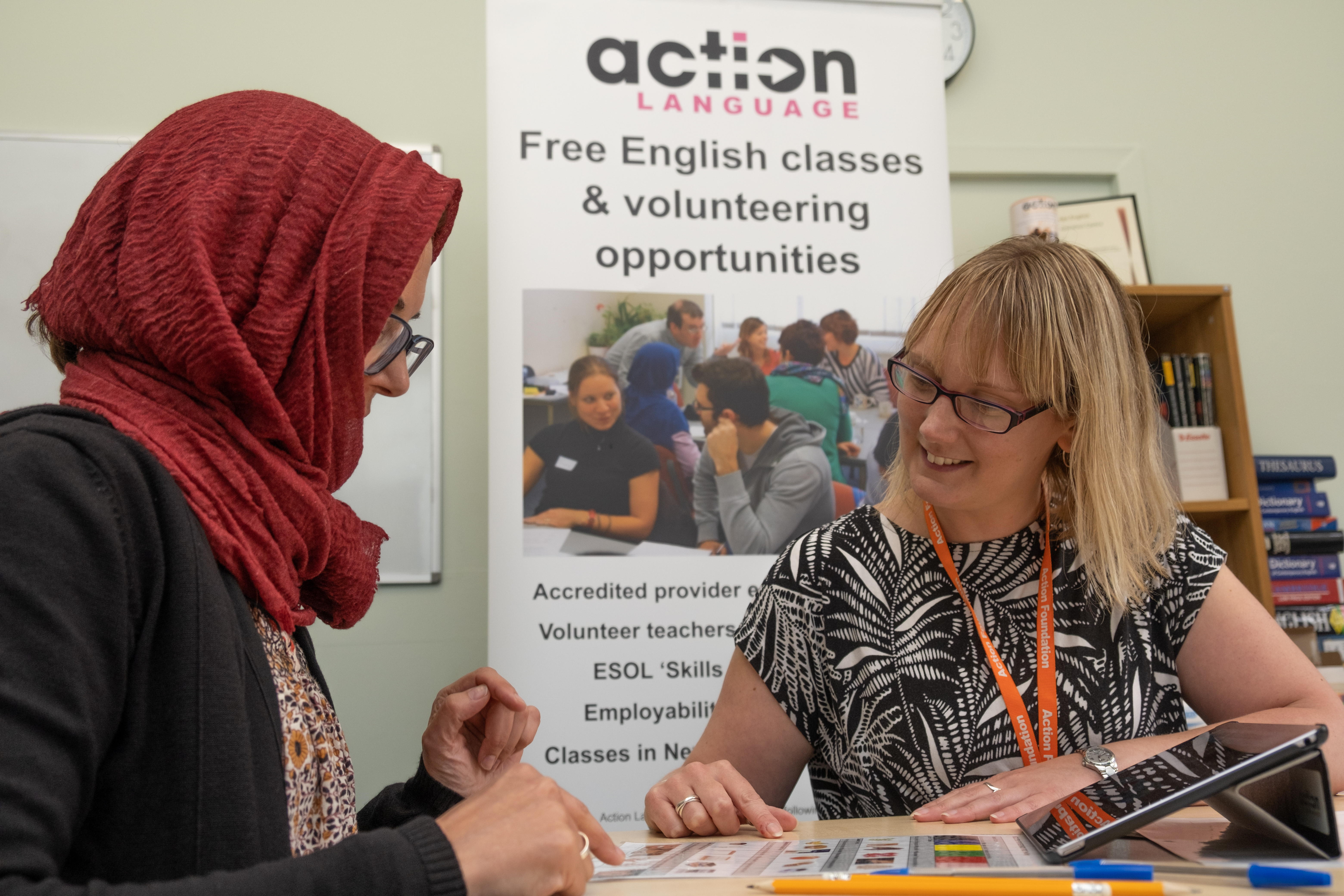 Two women sitting at a table pointing to some papers. A banner advertising English Classes is in the background