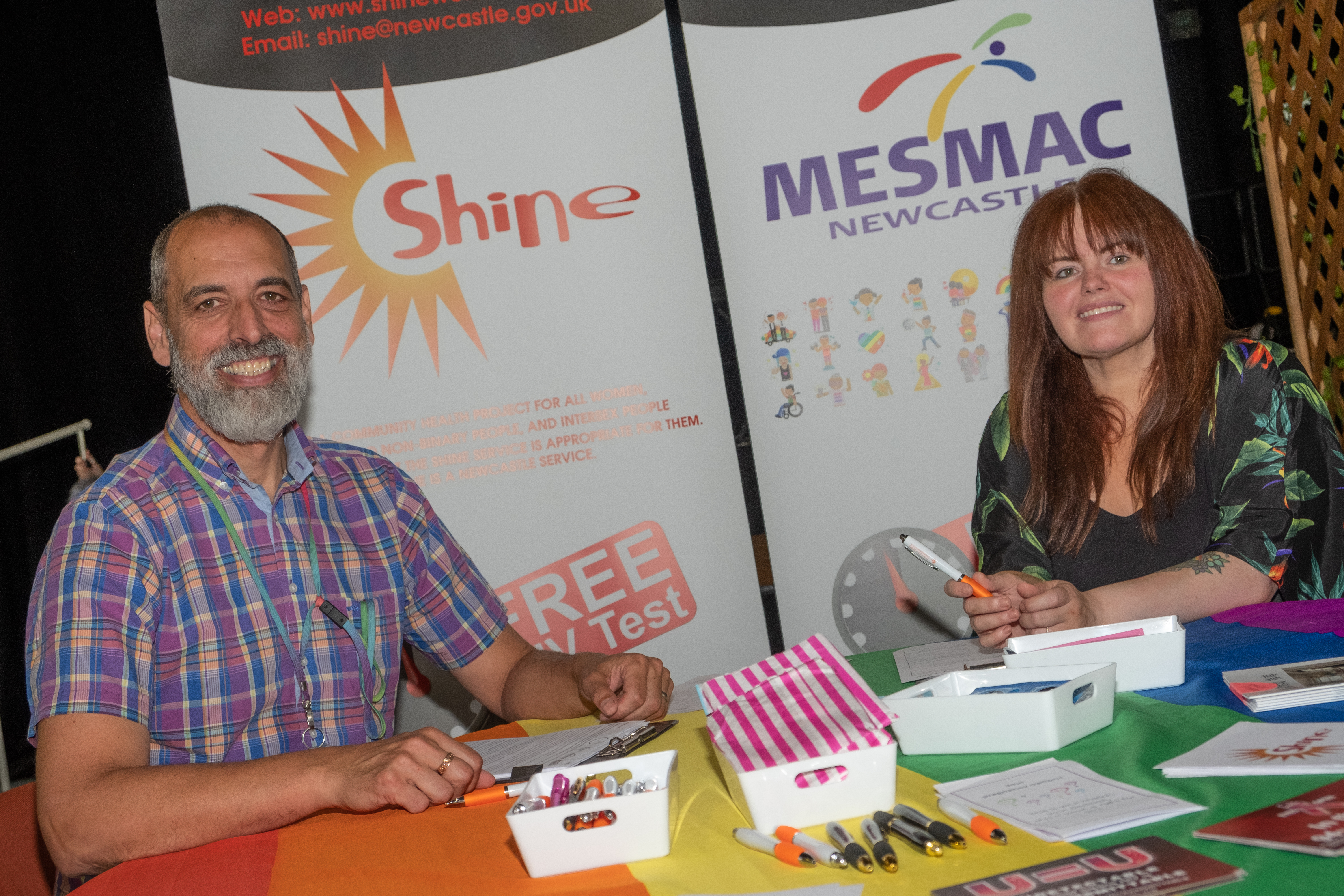 two people sit at a table with a rainbow flag table cloth and banners saying Shine and Mesmac behind them