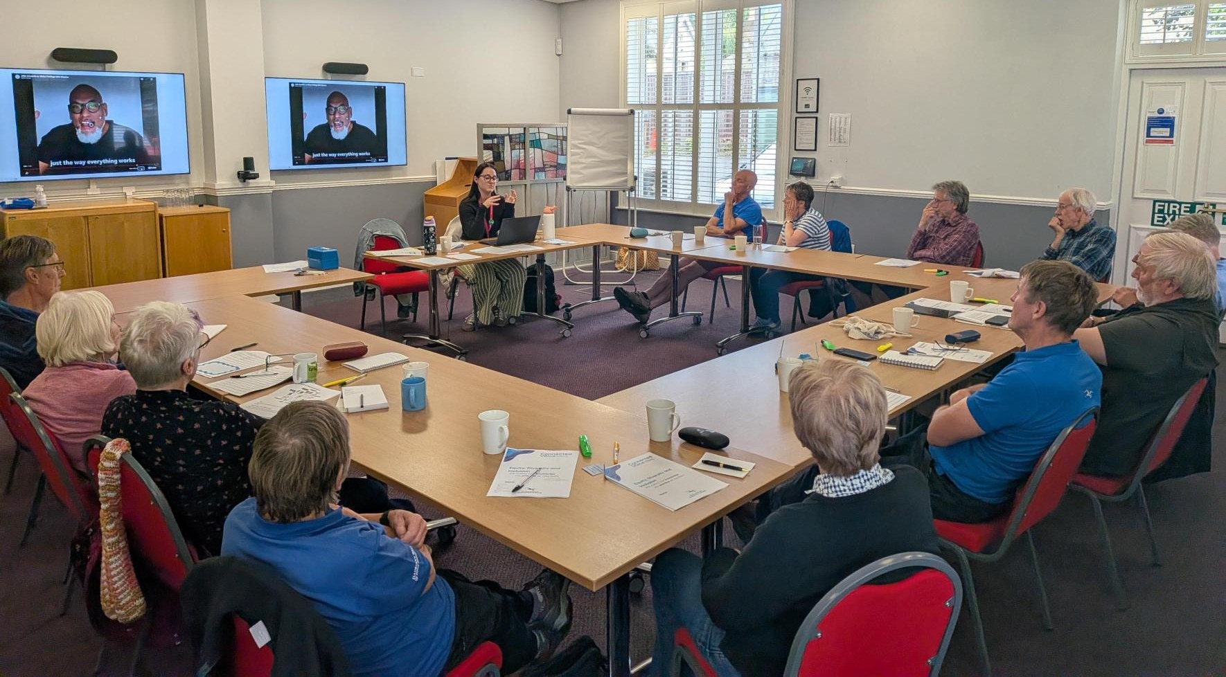 Attendees sit around a square table at a bespoke training session. A video plays on two screens in the background.