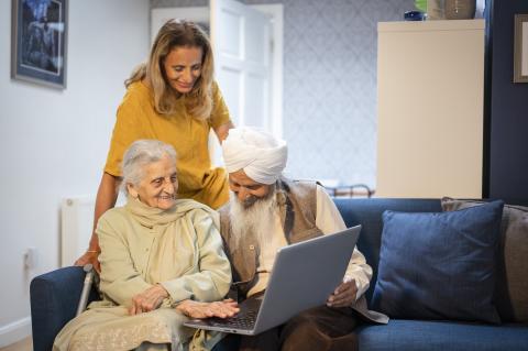 An older sikh man and woman sit on the sofa with a laptop and a keyworker overlooks helping them
